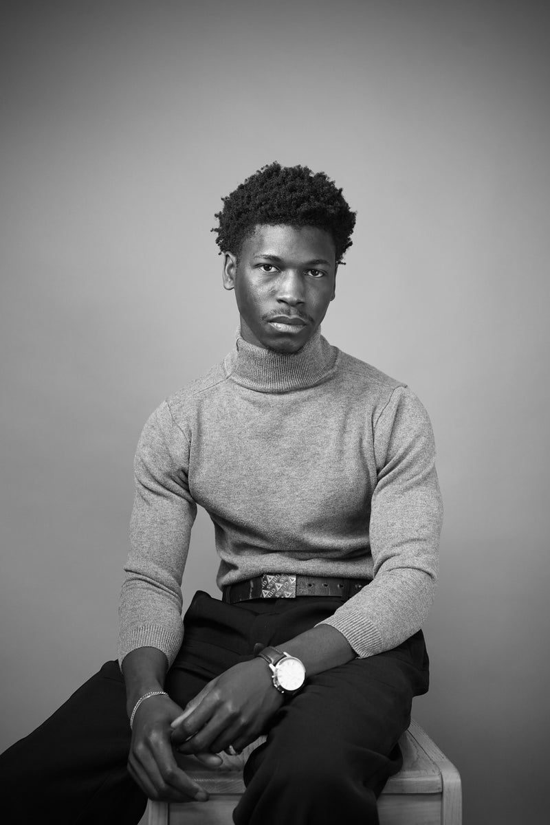 Man wearing a gray turtleneck sweater and dark pants against a plain background